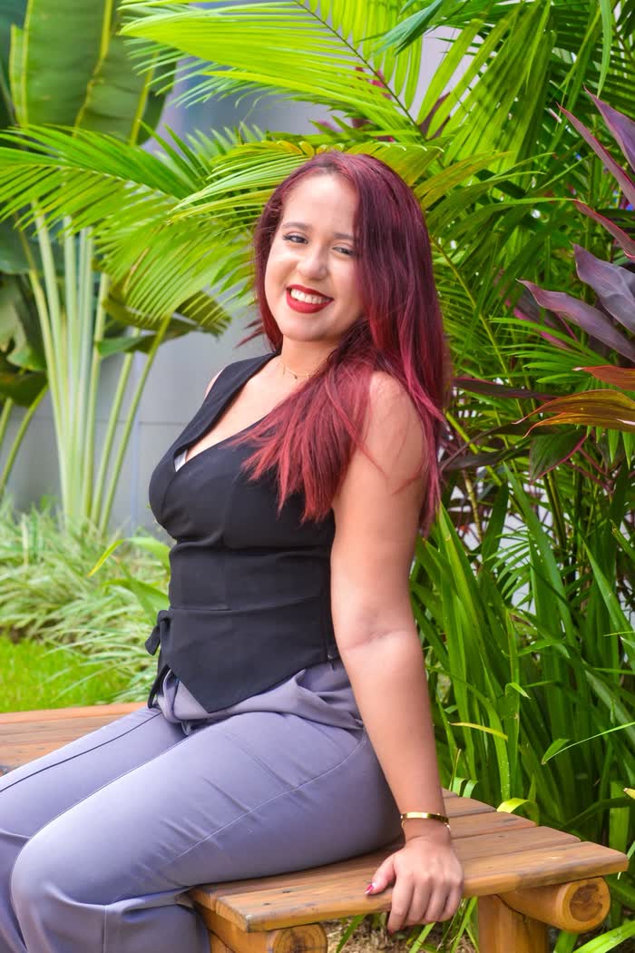 Retrato de Júlia sorrindo sentada em um banco de madeira em meio a plantas